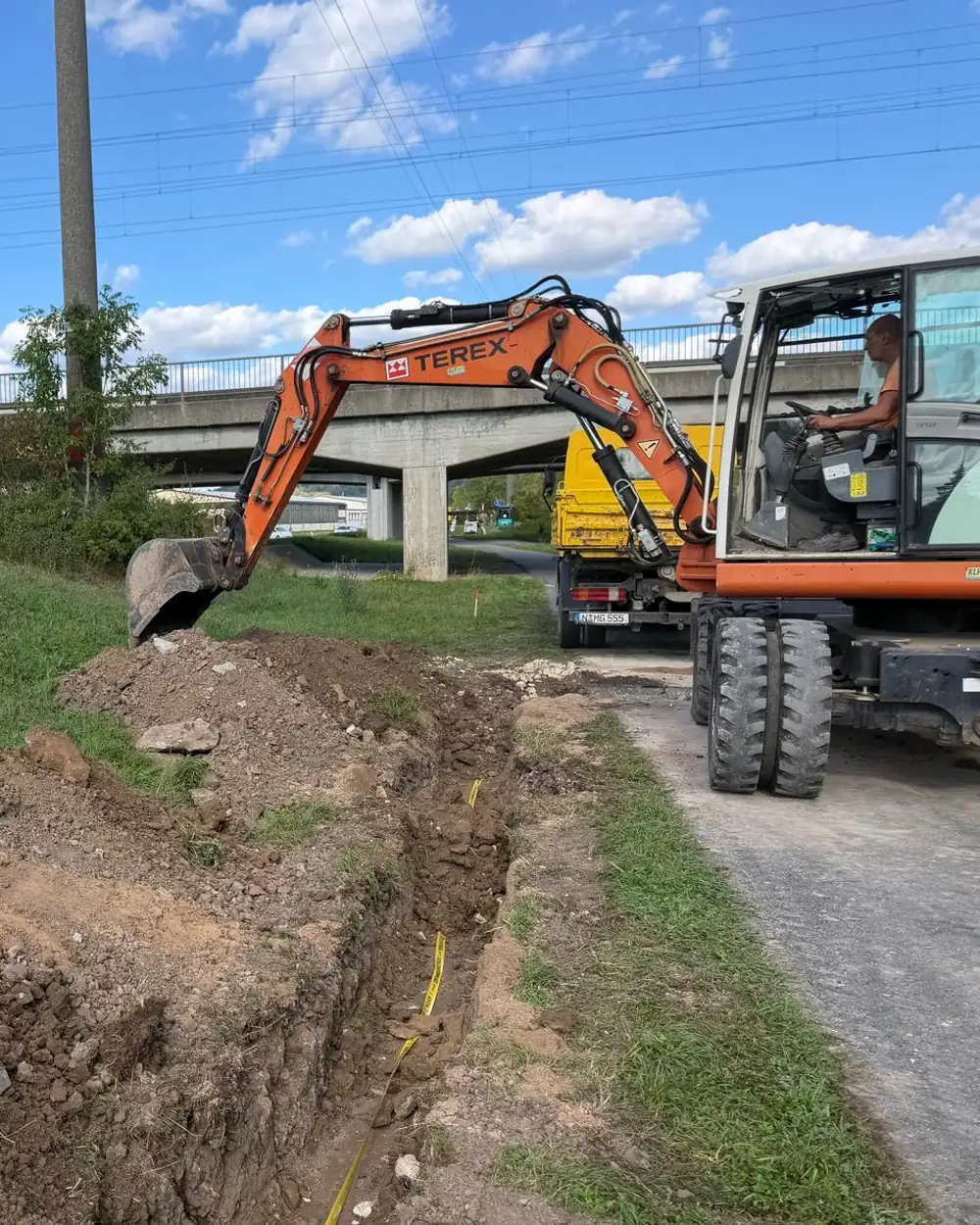 Mobilbagger im Einsatz am offenen Kabelgraben, Bahnstrecke im Hintergrund